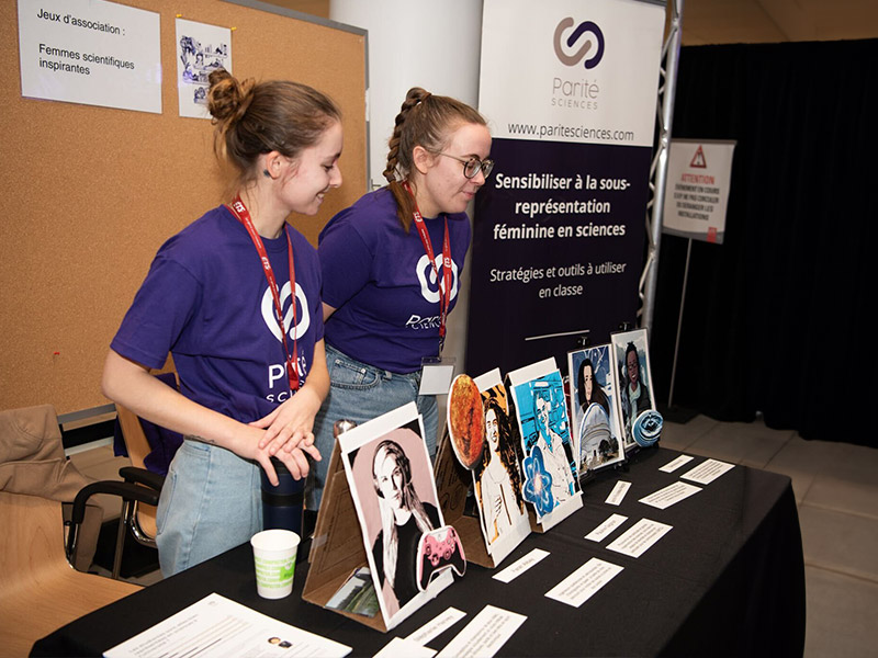 Two young girls in purple t-shirts and blue jeans are standing behind a table with a black tablecloth. On the table, there are 4 letter-sized posters standing on makeshift cardboard easels featuring famous women scientists.